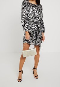 Abito a stampa leopardata bianco e nero con maniche lunghe, vita stretta e gonna fluida. Abbinato a una pochette metallica argento e tacchi neri.