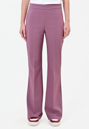 Donna che indossa pantaloni malva a vita alta e a gamba larga e sneakers rosse e bianche, in piedi su uno sfondo bianco uniforme.