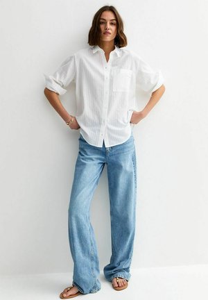 Femme aux cheveux bruns courts portant une chemise blanche à boutons avec les manches retroussées, un jean large bleu clair, des bracelets et des sandales.