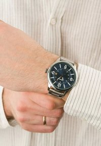 Montre en acier inoxydable avec un cadran bleu, grands chiffres blancs, affichage de la date et bracelet en maillons métalliques, portée sur une chemise à rayures claires.