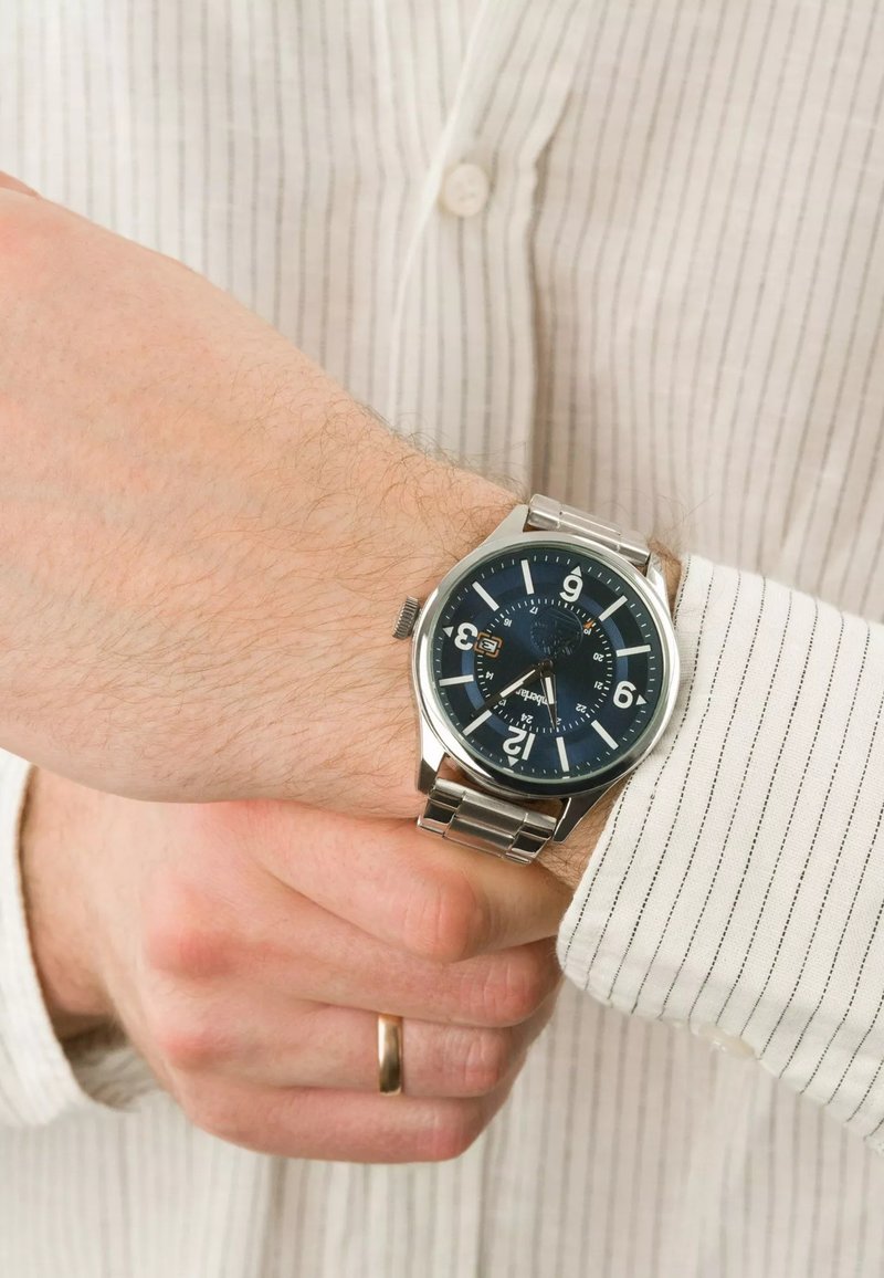 Montre en acier inoxydable avec un cadran bleu, grands chiffres blancs, affichage de la date et bracelet en maillons métalliques, portée sur une chemise à rayures claires.