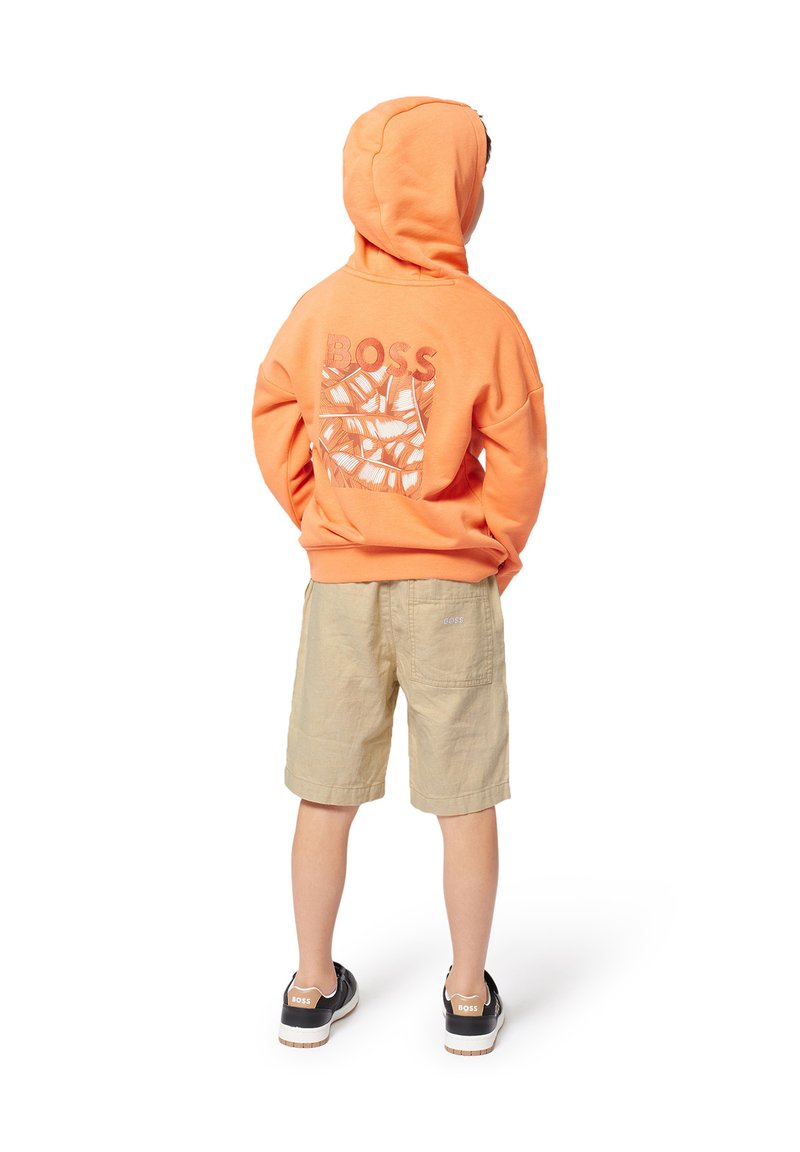 Hoodie orange avec le logo "BOSS" et un motif de feuilles au dos, associé à un short beige et des chaussures noires à semelles blanches.