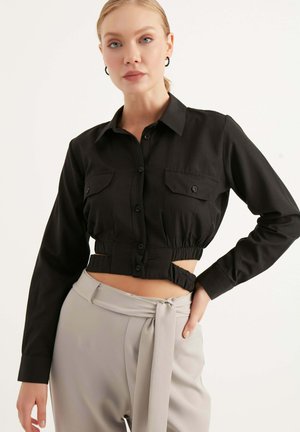 Camicia nera corta con bottoni e maniche lunghe, dotata di due tasche sul petto e con lati arricciati. Abbinata a pantaloni grigi chiari con cintura.