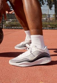 Sportssko i hvid med sorte accenter, der har en struktureret overdel og en tyk, polstret sål, designet til støtte under leg.