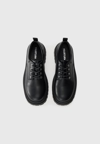 Zapatos de cuero negro con punta redonda, cierre de cordones y seis ojales metálicos. Suela de goma gruesa y una pestaña trasera para facilitar el calzado.