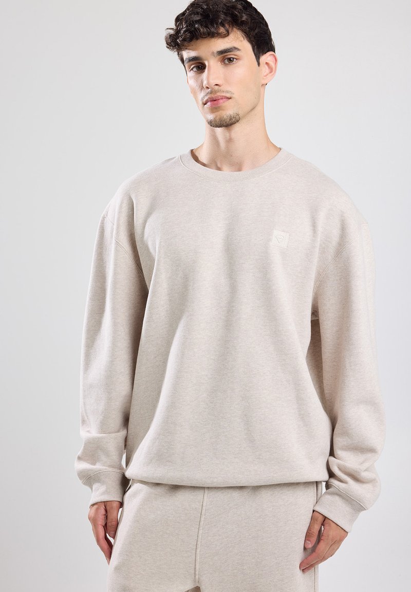 Felpa beige con collo a giro, maniche lunghe e una piccola toppa con logo sul petto. Realizzata in un tessuto morbido e texturizzato.