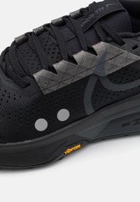 Sort sneakers med overdel i mesh, teksturerede grå detaljer, to refleksdæmpende prikker og en Vibram ydersål. Letvægtsdesign, der er velegnet til stier.