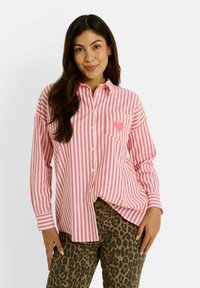 Camisa de botones a rayas en rosa y blanco con un bolsillo en el pecho. Llevada con pantalones de leopardo marrón. Mangas largas y corte holgado.