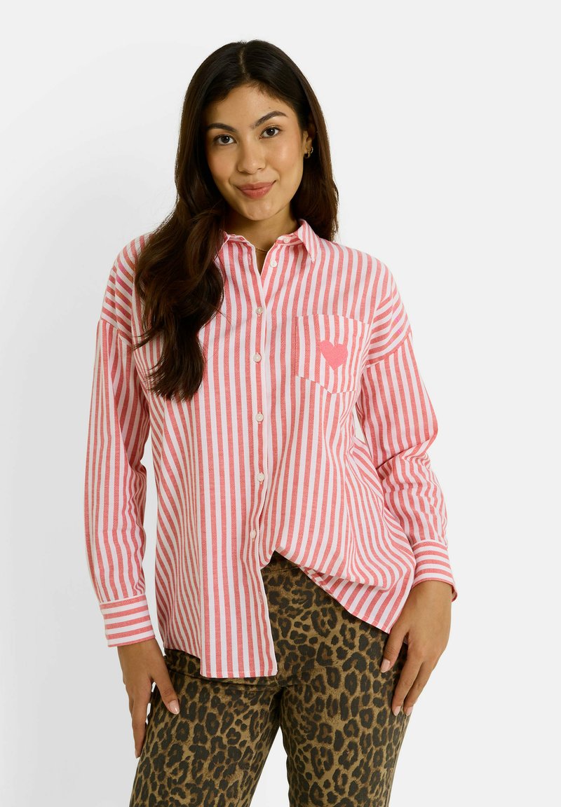 Camisa de botones a rayas en rosa y blanco con un bolsillo en el pecho. Llevada con pantalones de leopardo marrón. Mangas largas y corte holgado.