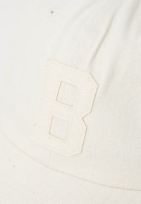 Cream-colored cap made of textured fabric, featuring a prominent embroidered letter "B" on the front. Simple, clean design with no patterns.