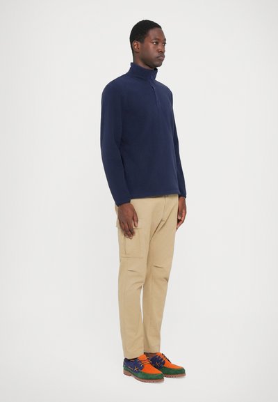 Marineblå fleecepullover med lynlås-hals, parret med beige cargo bukser og multikolore sko med grønne, orange og blå detaljer.