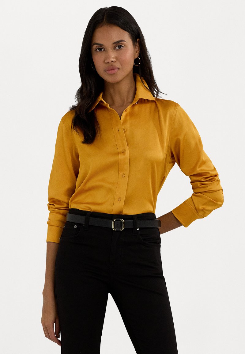 Camisa de botão de manga longa amarelo mostarda com colarinho estruturado e tecido suavemente texturizado, combinada com jeans pretos e um cinto justo.