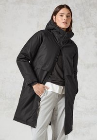 Zwarte parka-jack met capuchon, voorzien van een rits aan de voorkant, lange mouwen en een losse pasvorm. Draagt over een grijze trui en sweatpants.