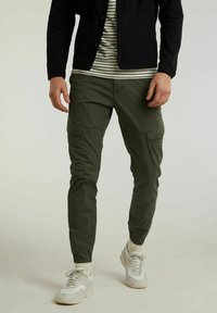 Hombre con pantalones cargo verde oliva, zapatillas blancas, camisa a rayas y chaqueta negra de pie sobre un fondo claro.