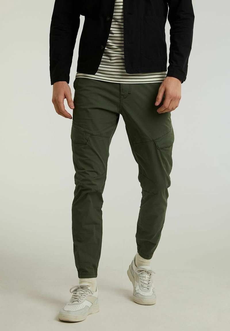 Hombre con pantalones cargo verde oliva, zapatillas blancas, camisa a rayas y chaqueta negra de pie sobre un fondo claro.