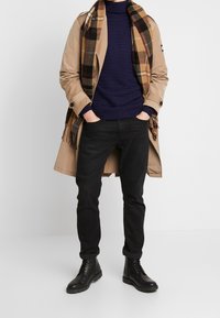 Beżowy trencz na niebieskim swetrze o fakturze, zestawiony z czarnymi dżinsami i czarnymi butami na sznurowadła. Uzupełniony brązowym szalikiem w kratkę.