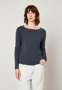 Ilgas rankovės tamsiai mėlynas marškinėliai iš tekstūruotos medžiagos su plačiu iškirpimu, laisva forma ir tiesiu apvadu, derinami su šviesiomis džinsinėmis kelnėmis.