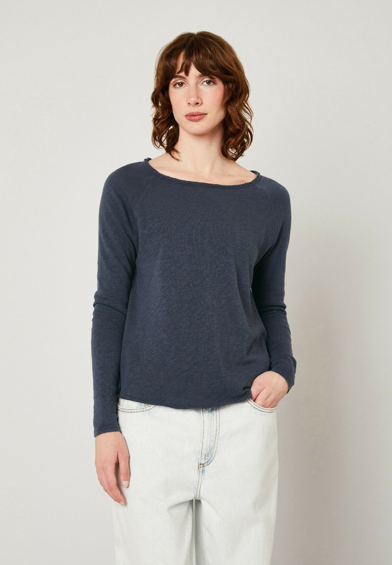 Ilgas rankovės tamsiai mėlynas marškinėliai iš tekstūruotos medžiagos su plačiu iškirpimu, laisva forma ir tiesiu apvadu, derinami su šviesiomis džinsinėmis kelnėmis.