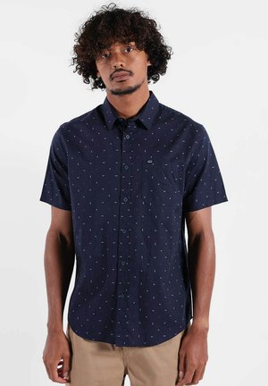 Homme aux cheveux bouclés portant une chemise marine à manches courtes à boutons avec petits motifs blancs et un pantalon beige devant un fond blanc.