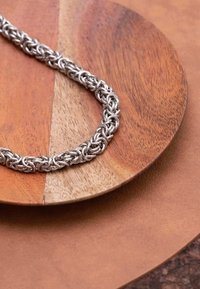 Zilveren ketting met complexe onderling verbonden schakels, geplaatst op een ronde houten schotel met contrasterende texturen en tinten.