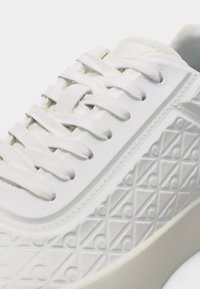 Κοντινή λήψη λευκού αθλητικού παπουτσιού με ανάγλυφο γεωμετρικό σχέδιο και λευκά κορδόνια σε ανοιχτόχρωμη σόλα.