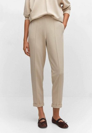 Pantalon beige taille haute avec ourlets roulés porté avec des ballerines marron foncé sur une personne debout devant un fond uni.