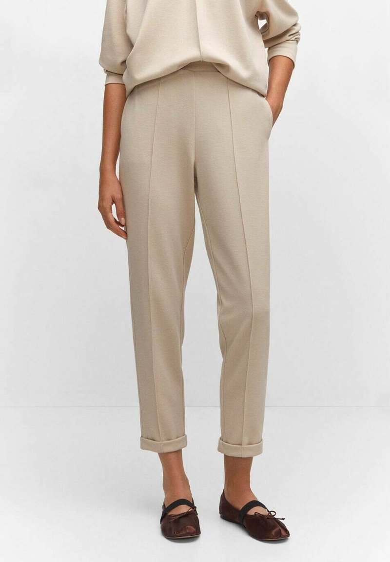 Pantalon beige taille haute avec ourlets roulés porté avec des ballerines marron foncé sur une personne debout devant un fond uni.