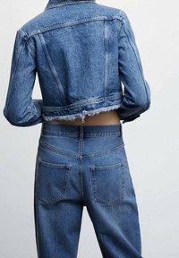 Takana näkymä henkilöstä, joka pukeutuu lyhennettyyn siniseen farkkutakkiin, jossa on purjeet, ja korkeavyötäröisiin sinisiin farkkuihin.