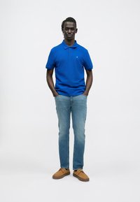 Férfi áll, kezét a zsebében tartja, kék galléros pólót, világoskék farmert és barna cipőt visel, egyszerű fehér háttér előtt.