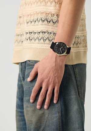 Schwarze Uhr mit goldenen Akzenten, runder Ziffernblatt, römische Ziffern, Silikonarmband, getragen an einer Hand gegen ein helles Strickoberteil und Jeans.
