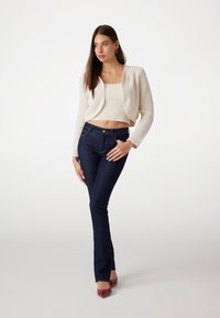 Lichtcrème gebreide cardigan over een bijpassende cropped top, gecombineerd met donkerblauwe flared jeans en rode puntige schoenen. Eenvoudige ontwerpdetaillering.
