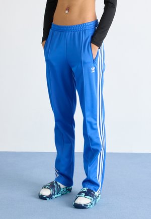 Persoon die blauwe Adidas trainingsbroek met witte strepen, zwarte crop top met lange mouwen en blauw patroon slip-ons draagt en op een blauwe vloer staat.