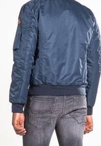 Personne portant une veste bomber bleue et un jean gris, vue de dos avec les mains détendues le long du corps.