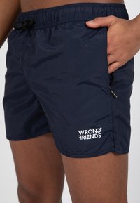 Námořnické modré plavky vyrobené z lehkého materiálu. Přednosti zahrnují elastický pas se šňůrkou a bílé logo na levé nohavici.