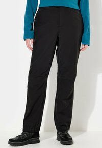 Black waterproof pants with a straight cut, elastic cuffs, and side pockets, paired with black rugged sneakers and a blue long-sleeve top.