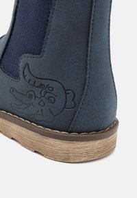 Närbild av en marinblå stövel med elastisk sidopanel, ljusbrun texturerad sula och präglat tecknat krokodilansikte på hälområdet.