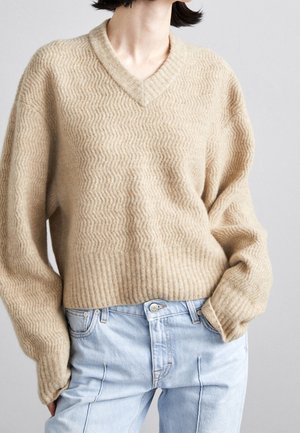 Beżowy sweter z fakturą, dekoltem w serek oraz prążkowanymi ściągaczami na mankietach i dole, noszony z jasnoniebieskimi jeansami z wysokim stanem, ręce w kieszeniach, na jednolitym tle.