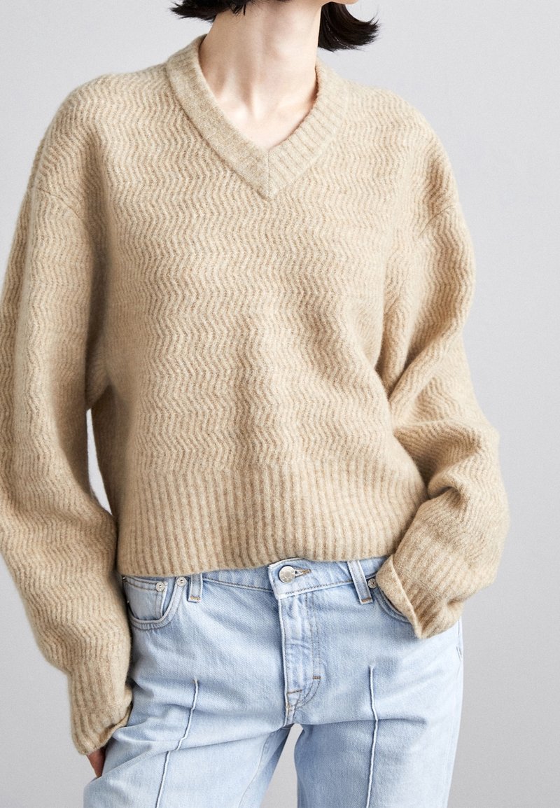 Pull beige texturé à col en V avec poignets et ourlet côtelés, porté avec un jean taille haute bleu clair, mains dans les poches, sur fond uni.