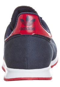 Laivastonsininen urheilujalkine punaisilla yksityiskohdilla, jossa on sileä pinta ja valkoinen pohja. Takana on selvästi näkyvissä Adidas-logot.