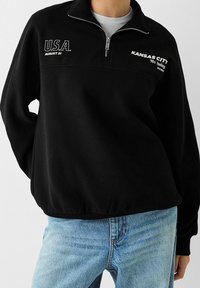 Pullover in pile nero con colletto zip, con testo bianco: "U.S.A." e "KANSAS CITY anni '90". Orlo e polsini a coste; vestibilità morbida.