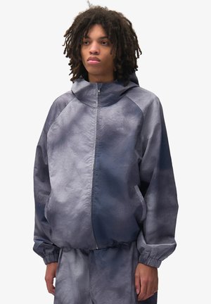 Giovane con capelli ricci scuri indossa una giacca con cappuccio blu-grigio larga e pantaloni abbinati, in piedi davanti a uno sfondo chiaro e uniforme.