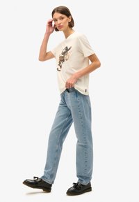 T-shirt color crema con stampa grafica, abbinata a jeans chiari a vestibilità ampia e mocassini neri. Design semplice e informale.