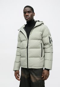 Vaaleanharmaa puuvilla-puffa-takki hupulla, jossa on edessä vetoketju, kaksi sivutaskua ja yksi käsivartetasku mustilla yksityiskohdilla ja mattapinnalla.