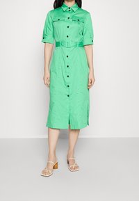 Grünes Kurzarm-Kleid mit Knopfleiste, taillierter Taille, seitlichen Schlitzen und schwarzen Knöpfen. Kombiniert mit beige Blockabsatzsandalen. Glatte Textur.