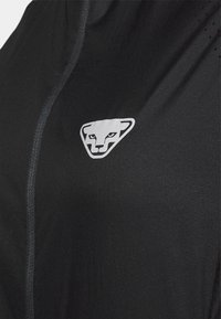 Haut de sport noir à texture lisse, avec une fermeture éclair grise et un logo triangulaire blanc ressemblant à un visage d'animal stylisé.