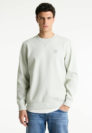 Felpa verde chiaro con texture e collo a giro, caratterizzata da un piccolo logo sul petto e polsini e fondo a costine. Indossata con jeans blu.