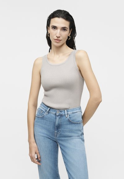 Γυναίκα με μακριά σκούρα μαλλιά που φοράει χρυσούς κρίκους σκουλαρίκια, γκρι αμάνικη ριμπ κορυφή και μπλε ψηλόμεσα τζιν, σε λευκό φόντο.