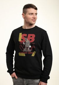 Černá mikina s kulatým výstřihem, na které je grafický design kreslené postavičky na skateboardu, doplněná žlutou páskou a výrazným písmem.