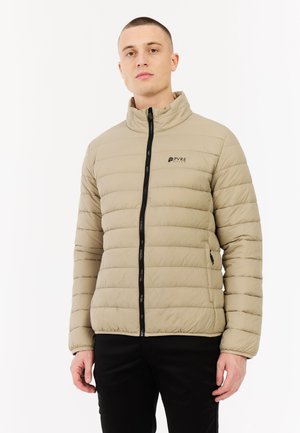 Man in een beige gewatteerde jas met zwarte rits en logo, staand tegen een eenvoudige witte achtergrond.