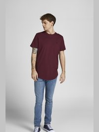 Burgunderfarbendes T-Shirt mit kurzen Ärmeln und abgerundetem Saum, kombiniert mit hellblauen Slim-Fit-Jeans und navyfarbenen Sneakern. Hat Tattoos am Arm.